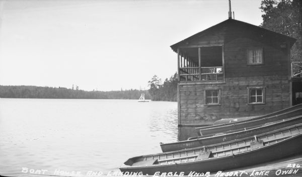 View along shoreline of wooden rowboats pulled up at the landing, and a boathouse with a second-story porch at the Eagle Knob Resort on Lake Owen. There is a diving dock in the background floating in the lake. Caption reads: "Boat House and Landing, Eagle Knob Resort — Lake Owen."