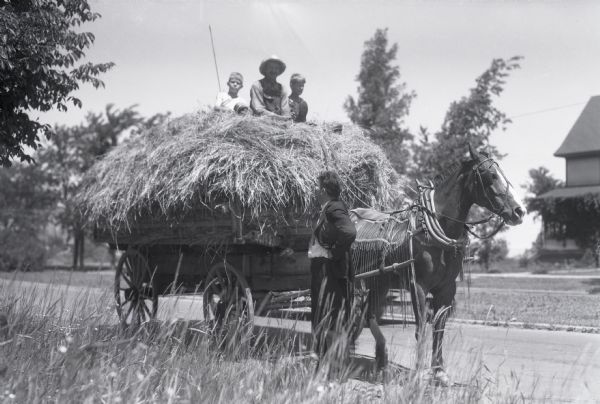 View of two boys and a man sitting on top of a loaded hay wagon hitched up to one horse wearing a fly-net and blinders. A man in a suit is standing next to the horse. Trees and a house are in the background.
