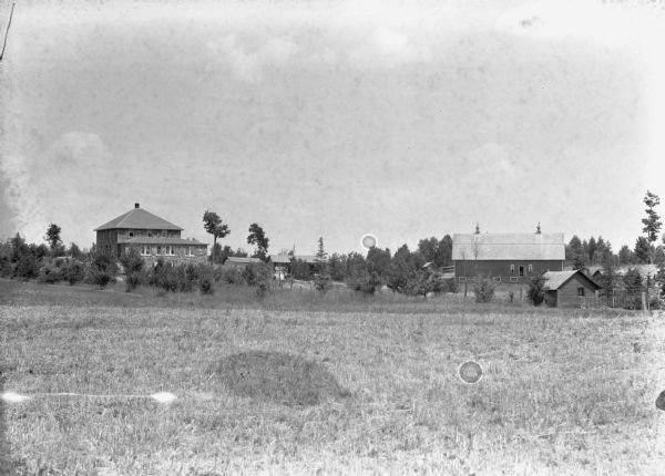 View across field overlooking an unidentified farm. On the left is a two-story stone building, possibly the farmhouse. On the right is a barn. Other outbuildings are among trees.