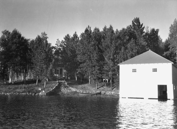 View across water of a shoreline with stone steps and wall leading up to a cabin among trees. There is a dog on the left near the steps, and a wooden canoe pulled up on shore. On the right is a white two-story boathouse. The cabin and lake are unidentified.