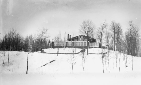 Exterior view of the front facade of a large two-story cabin with two chimneys. There are trees around the cabin, and snow covering the ground. A birdhouse is visible in the foreground on the right.