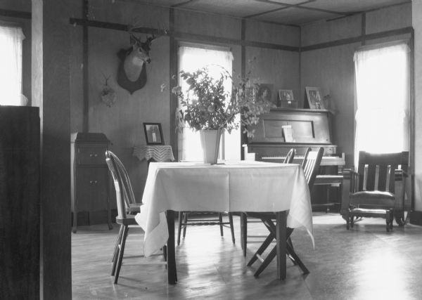 Interior view of a room with a dining set of table and chairs. On the wall on the left is what appears to be a Victrola cabinet. On the right is a piano, rocking chair and stool. There is a flower arrangement on the table, photographs on top of the piano, and a buck head mount is displayed on the wall near a wasp's nest.