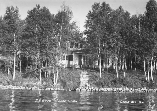 View over water of the shoreline, stone steps, and the A.F. Boyd cabin or lake home amongst trees.