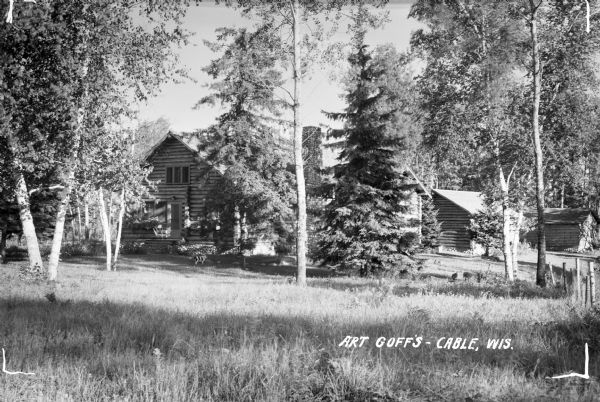 View across lawn of Art Goff's log cabin with stone chimney and foundation. Two other log outbuildings are on the far right. There is a black and white dog standing near the cabin.