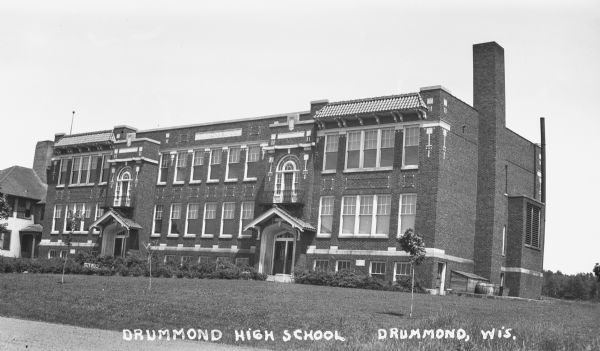 Exterior view of a two-story brick building, the Drummond High School. There is some decoration around the windows, front doors, and in the brickwork. The old elementary school is in the background on the far left.<p>“The Drummond State Graded – High School. In the summer of 1922, construction was started on a new two-story brick school. This was to be a combination grade and a full four year high school.  … Some difficulty was experienced in digging the basement, as the soil was all red clay and the hole kept filling with water. Construction was carried on throughout the summer and fall and into the winter. … The contractor was the Weaver Construction Company of Owen, Wisconsin.   Once completed, the school had a fairly large gym, boys and girls shower rooms, a lunch room, a kitchen and one class room in the lower level. … On the first floor there were six classrooms and boys and girls rest rooms.  On the second floor were four class rooms, a large assembly hall with library and two office rooms.  On the fourth of September, 1923, the school opened its doors for the first time.”  - Information from the "Drummond Centennial 1882-1982," 2nd printing, by Gordon G. Sorenson, pages 255-256.</p>