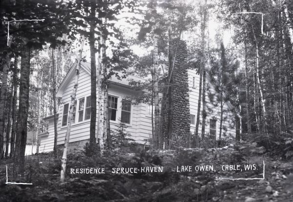 Exterior view of a one-story house with a tall stone chimney. Labeled as Residence Spruce-Haven on Lake Owen. The house is surrounded by trees.