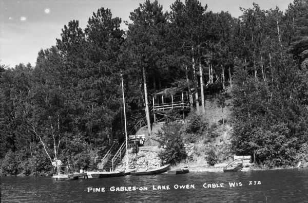 View from lake of a few boys on a dock as well as four wooden boats pulled up along the dock or the shoreline. There is a diving platform at the end of the pier. There are steep wooden stairs leading up the hill on the shore, which has a large roofed landing with benches half way up. There are many pine trees and shrubs on the hill.