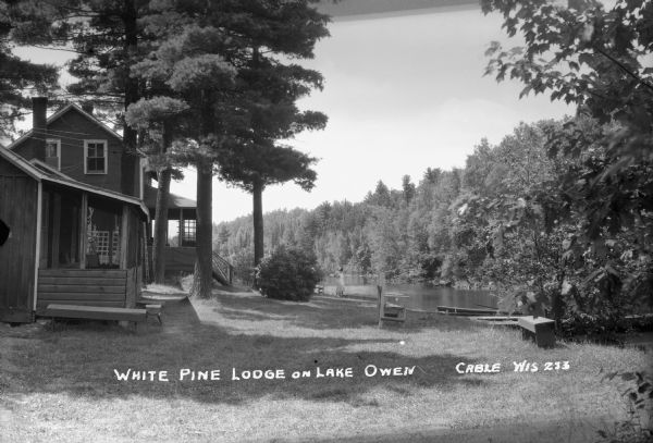 Two women stand on the lawn looking at Lake Owen. There are wooden benches, trees, and a rowboat on the right. The White Pine Lodge with porches is on the left.