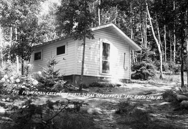 Exterior view of a small one-story cottage surrounded by trees and flowering plants. There is a large fishing hook on the wall near the door. There is a dirt path in front of the building. The negative is identified as "The Fisherman Cottage - Private Slacks 'Beautyrest' Lake Owen."