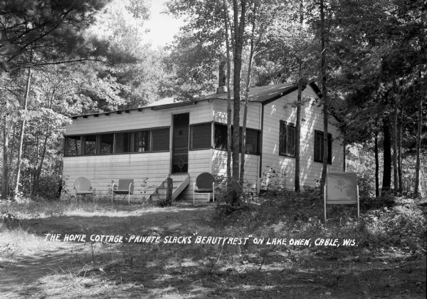 Exterior view of a small one-story cabin with a front porch. There are four classic metal lawn chairs outside the cottage which is surrounded by trees. The negative is labeled as "The Home Cottage - Private Slacks 'Beauty Rest' on Lake Owen."