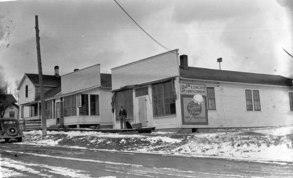 View across road of a man and a dog standing on the front stoop of a one-story building that has a sign that reads “Art’s Lunches and Confectionery, Ask for Velvet Ice Cream, New Quick Freeze.” The next building to the left has a sign that says “Art’s Tavern.” The third building is a two-story wooden house with a front porch. There is snow on the ground and one automobile on the far left, parked in front of the tavern.