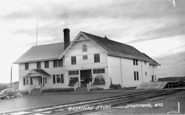 Exterior three-quarter length view of a large two-story building with one chimney. There are two main entrances with steps leading up to the doorways of what is labeled the Drummond Store. There is one automobile parked in front. In the foreground are railroad tracks, and on the right side of the building is a wooden platform or loading dock between the building and the railroad tracks.