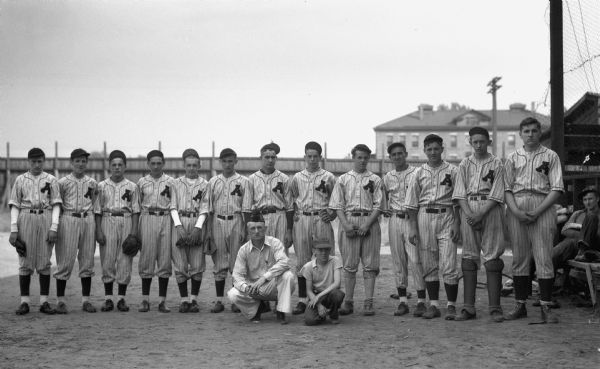 Group portrait outdoors of thirteen boys standing in a line wearing baseball hats and baseball uniforms with a large "A" on their shirts. One man and a younger boy are crouched down in front of the team, and another man is sitting on a bench on the far right. In the background is a wooden fence, and beyond a two-story brick building.