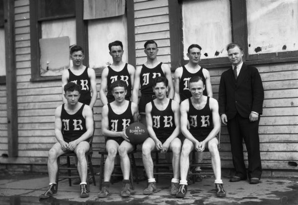 Group portrait of a basketball team showing eight men wearing basketball uniforms with the letters "IR" on their shirts, and one man on the right wearing a suit. In the center a man holds a basketball that says "I.R. Bearcats 1932-1933." The men are posing outdoors in front of a wood sided building.