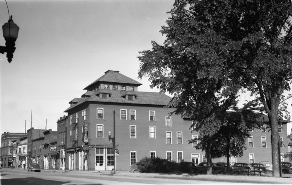 Exterior view of Hotel Menard, a three-story building with what appears to be brick and wood siding. Looking down Main Street West, what used to be called Second Street West, the front facades of the other buildings and storefronts are visible. Signs on the buildings read "Hotel Menard," "Bus Depot, Washburn, Bayfield," "Menard Coffee Shop," "Grill," and "Schlitz on draught." There is one automobile parked on Main Street and more are parked along the road on the right, Chapple Avenue.<p>Hotel Menard was located at 701 Second Street West (today this would be the corner of Main Street West and Chapple Avenue) and is listed in the Ashland City Directory from 1926 to 1956.