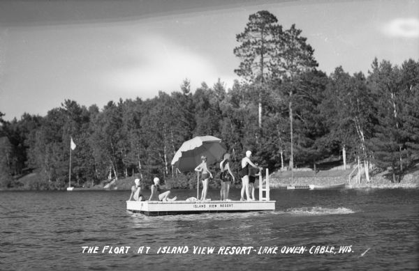 View across water of seven people wearing bathing suits on the float or wooden raft in Lake Owen at the Island View Resort. There is a large umbrella on the float, which has the words "Island View Resort" painted on the side. In the background is the wooded shoreline with two docks, boats, and a flag on a flagpole.