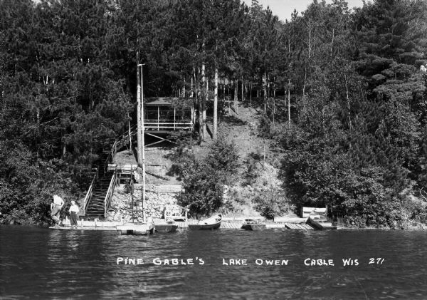 View from the water of three boys on the dock at the foot of a steep stairway. There is a large roofed landing with benches half way up the hill, and many pine trees and shrubs. Four wooden boats are pulled up on a wooden platform along the shoreline. A diving platform is on the end of the pier. The image is labeled as "Pine Gable's Lake Owen".