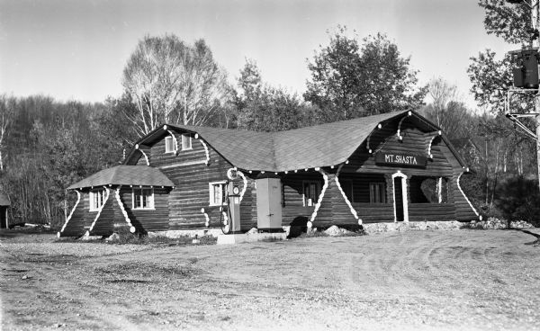 Exterior view of a large one-story log building that has a sign that says Mt. Shasta, located in the Upper Peninsula. There is a gravel drive and a gas pump and sign that says "Phillips 66."