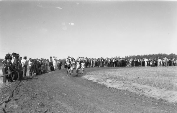 Crowd of spectators standing around a dirt race track while five boys race around the track.