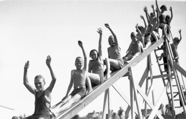 View of nine children wearing bathing suits waving at the camera man as they go down the swimming slide. Five more children wearing bathing suits are going up the ladder for the slide.
