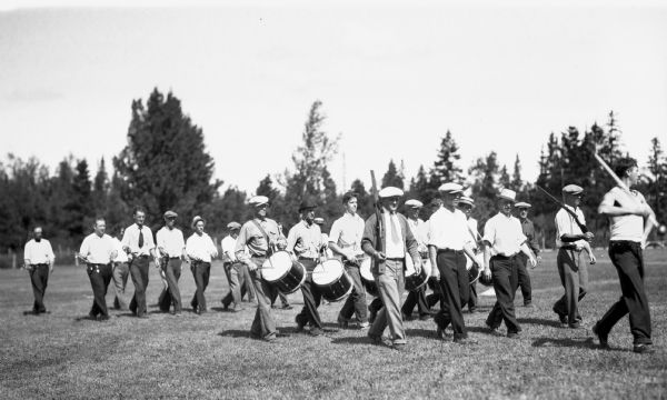 View of twenty-one men marching outside holding rifles, drums and bugles. Many of the men are wearing hats.