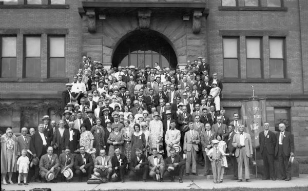 Large group portrait of the Wisconsin Rural Mail Carriers Association showing men, women, and a few children on the front steps of, and in front of, a brick and brownstone building.  The women are wearing dresses and hats, and the men have on suits and ties. A large banner on the far right reads: "Wisconsin Rural Letter Carriers Association."