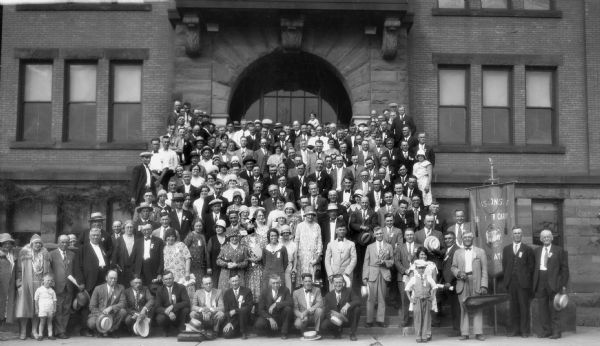 Large group portrait of the Wisconsin Rural Mail Carriers Association showing men, women, and a few children on the front steps of, and in front of, a brick and brownstone building. The women are wearing dresses and hats, and the men have on suits and ties. A large banner on the far right reads: "Wisconsin Rural Letter Carriers Association."