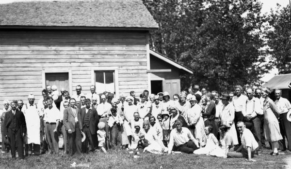 Large group portrait of men, women, and children in front of a wooden building. Some of the men are wearing suits and ties, and some of the women are wearing hats.