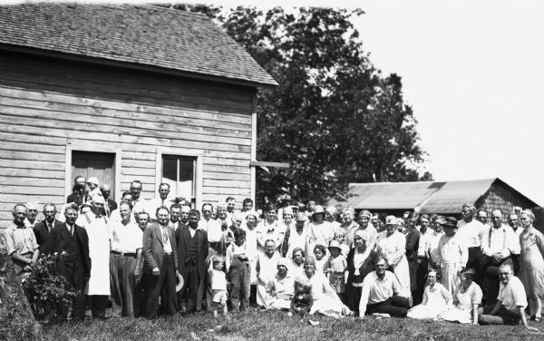 Large group portrait of men, women, and children out front of a wooden building. Some of the men are wearing suits and ties, and some of the women are wearing hats.