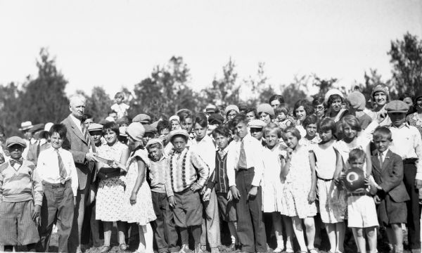 Girl wearing a hat and dress receiving an award from a man wearing a suit and tie while other children in a large crowd are around them.