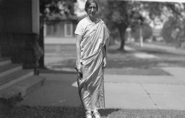 White woman standing outside on the sidewalk wearing a Sari, eyeglasses, and ballet shoes. There are buildings, sidewalks and trees in the background.