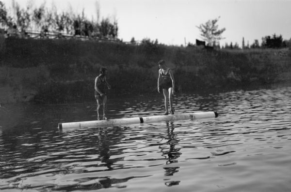 View of two men log rolling, standing on a log in the water. Both men are wearing bathing suits and tank tops. The young man on the right has "Eau Claire Wis" written on his tank top.