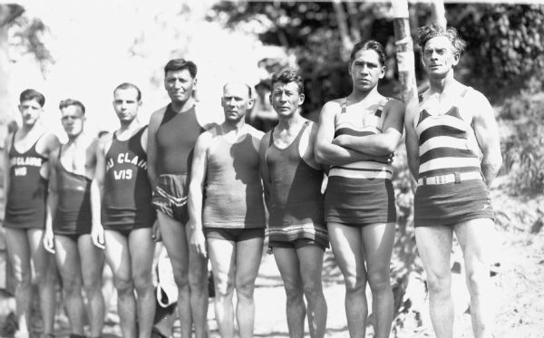 Group portrait of eight men standing in a line wearing bathing suits.  Two of the men have the words "Eau Claire Wis" on their tank tops. This is probably an outdoor group portrait of male log rolling contestants.