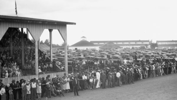 A large crowd of men, women, and children sit in the bleachers or stand along the edge of what appears to be the racetrack at the fair grounds. Behind the crowd there are many parked automobiles and a few buildings are visible in the distance.