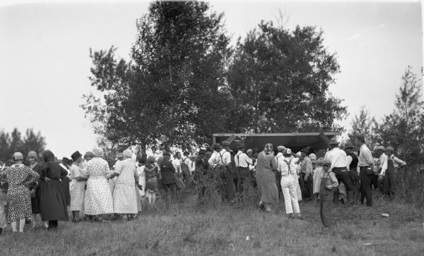 Men, women, and children in a crowd standing around outside talking in groups near a small outdoor shed. Most of the people have hats on. The women are wearing dresses, and many of the men are wearing suspenders. There are two boys in the foreground, one of whom is wearing a baseball hat with the 4-H clover symbol.