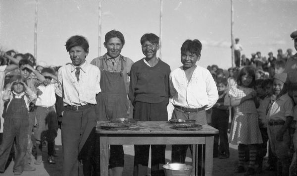Four boys smiling with pie filling on their faces standing around a table with four empty pie tins. Behind them is a large crowd of children.