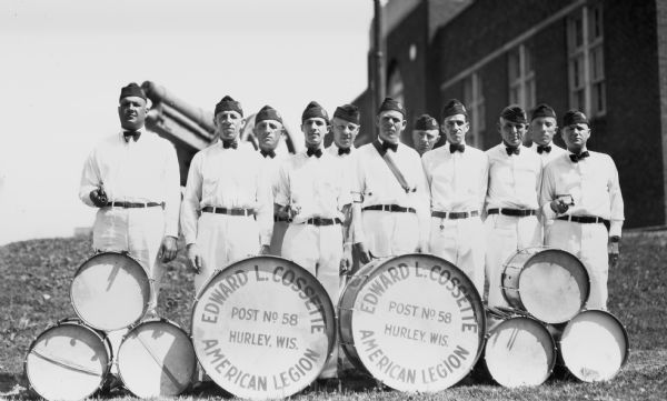 Group portrait of eleven men wearing hats, shirts and bow ties standing outdoors holding drum sticks behind eight drums. The two large drums in the center say “Edward L. Cossette, Post No. 58, Hurley, Wis. American Legion.” In the background is a building.