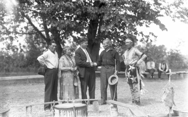Group portrait showing one Native American man shaking hands with John Chapple, while two other Native American men and one Native American woman wearing a decorated dress look on. In the foreground is a drum and benches, and in the background are people sitting on a bench and a tree.