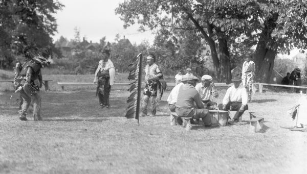 View of four men sitting outside around a drum while four other men wearing Native American clothing are dancing. In the background are two boys sitting on benches. Off to the far right adult and child sit under a tree.
