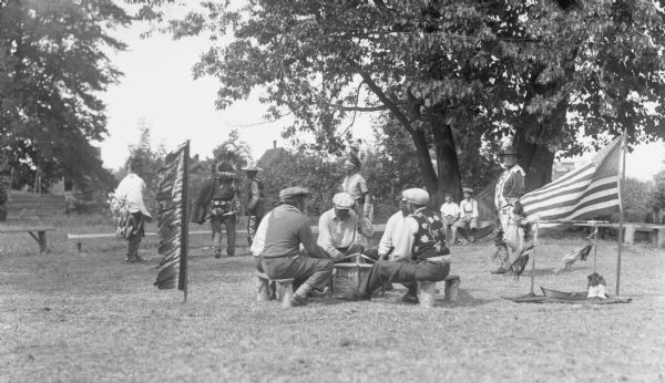 View of five men wearing hats sitting outside in a circle around a drum while five other men dressed in Native American clothing are standing behind the seated men. Two boys are sitting on benches in the background, with trees and shrubs visible behind them. Off to the right is an American flag and a Native American peace pipe.