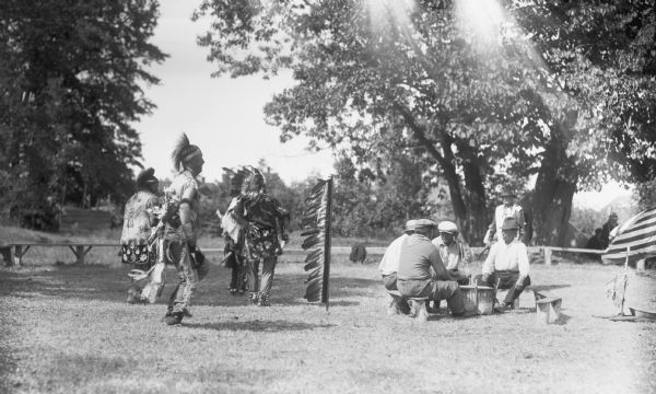 View of four men sitting outside around a drum while four other men wearing Native American clothing are standing off to the left. On the far right an American flag is visible. In the background are benches, a few other people, trees and shrubs.