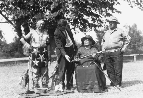 Three men standing outside, one is dressed wearing Native American clothing items, and another, wearing a suit and tie, is probably John Chapple. One elder woman wearing a dress and hat is sitting with her cane and holding a peace pipe.
