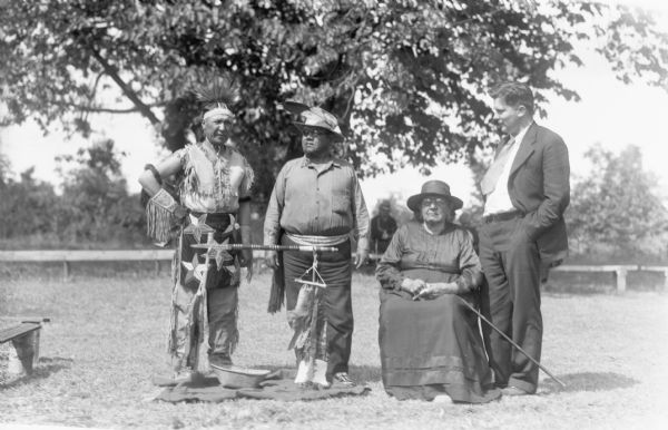 Three men standing outside, one is dressed wearing Native American clothing items, and another is wearing a suit and tie. One elder woman wearing a dress and hat is sitting holding her cane near the men.