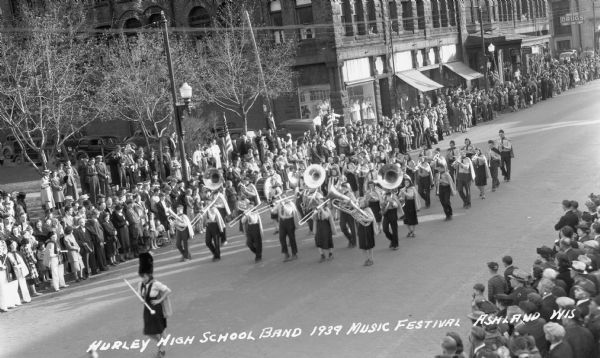 Elevated view of students from the Hurley High School Band marching in a parade at the Music Festival, on Main Street. Students are playing instruments including trombones, tubas, euphoniums, drums, trumpets, saxophones, and clarinets. Crowds of people watch from the sidewalks.