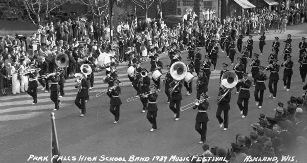 Elevated view of students from the Park Falls High School Band marching in a parade at the Music Festival, on Main Street.  Students are wearing marching band uniforms and hats, and are playing instruments including trombones, French horns, tubas, euphoniums, drums, trumpets, saxophones, and clarinets. Crowds of people watch from the sidewalks.