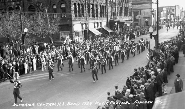 Elevated view of students from the Superior Central High School Band marching in a parade at the Music Festival, on Main Street. Students are wearing marching band uniforms and hats, and are playing instruments including trombones, tubas, drums, trumpets, saxophones, and flutes. Crowds of people watch from the sidewalks.