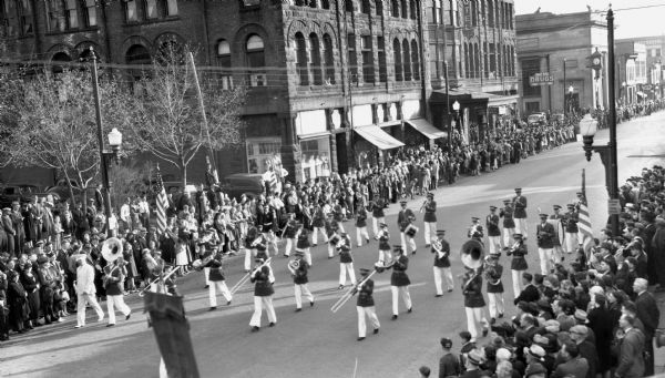 Elevated view of students from the Drummond High School Band marching in a parade at the Music Festival, on Main Street. Students are wearing marching band uniforms and hats, and are playing instruments including trombones, French horns, tubas, euphoniums, drums, trumpets, and saxophones. Crowds of people and American flags line the sidewalks.