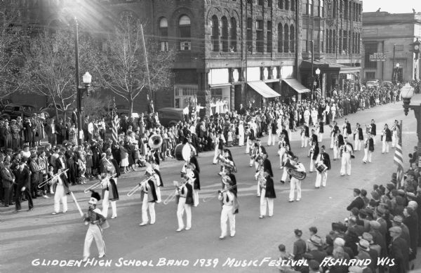 Elevated view of students from the Glidden High School Band marching in a parade at the Music Festival, on Main Street. Students are wearing marching band uniforms and hats, and are playing instruments including trombones, euphoniums, drums, trumpets, and flutes. Crowds of people watch from the sidewalks and the brownstone building across the street is the Knight Hotel.