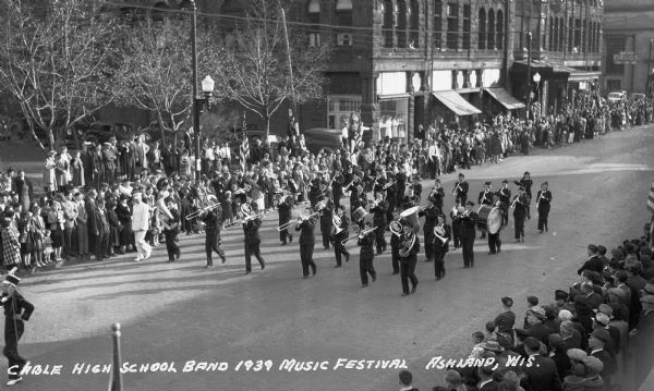 Elevated view of students from the Cable High School Band marching in a parade at the Music Festival, on Main Street. Students are wearing marching band uniforms and hats, and are playing instruments including trombones, French horns, tubas, drums, trumpets, clarinets, and flutes. Crowds of people watch from the sidewalks, and the Knight Hotel is across the street.