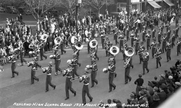 Elevated view of students from the Ashland High School Band marching in a parade at the Music Festival, on Main Street. Students are wearing marching band uniforms and hats, and are playing instruments including trombones, French horns, tubas, euphoniums, drums, trumpets, a bassoon, saxophones, clarinets, and flutes. Crowds of people watch from the sidewalks.